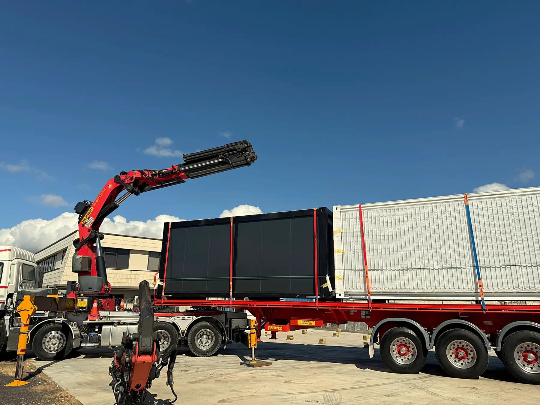 Folded expandable units strapped to a flatbed trailer beside a truck-mounted crane